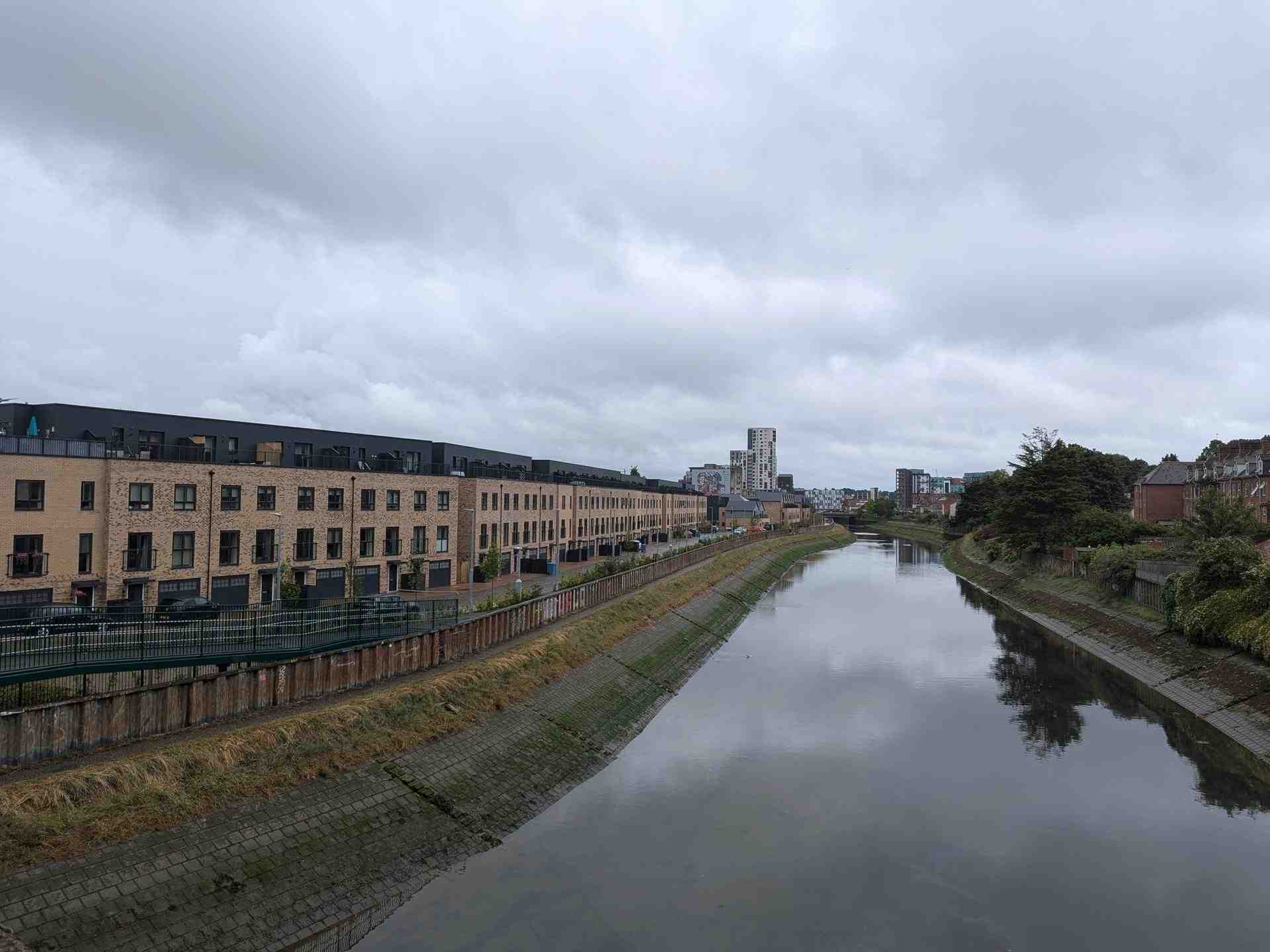 A grey river bounded by a man-made embankment of square stones, and a grassy verge above. On the left is a long row of new houses built with lightly-coloured bricks, with the top floor set back and clad in black. To the right, a long row of older houses built with darker bricks. In the distance, some high-rise buildings.