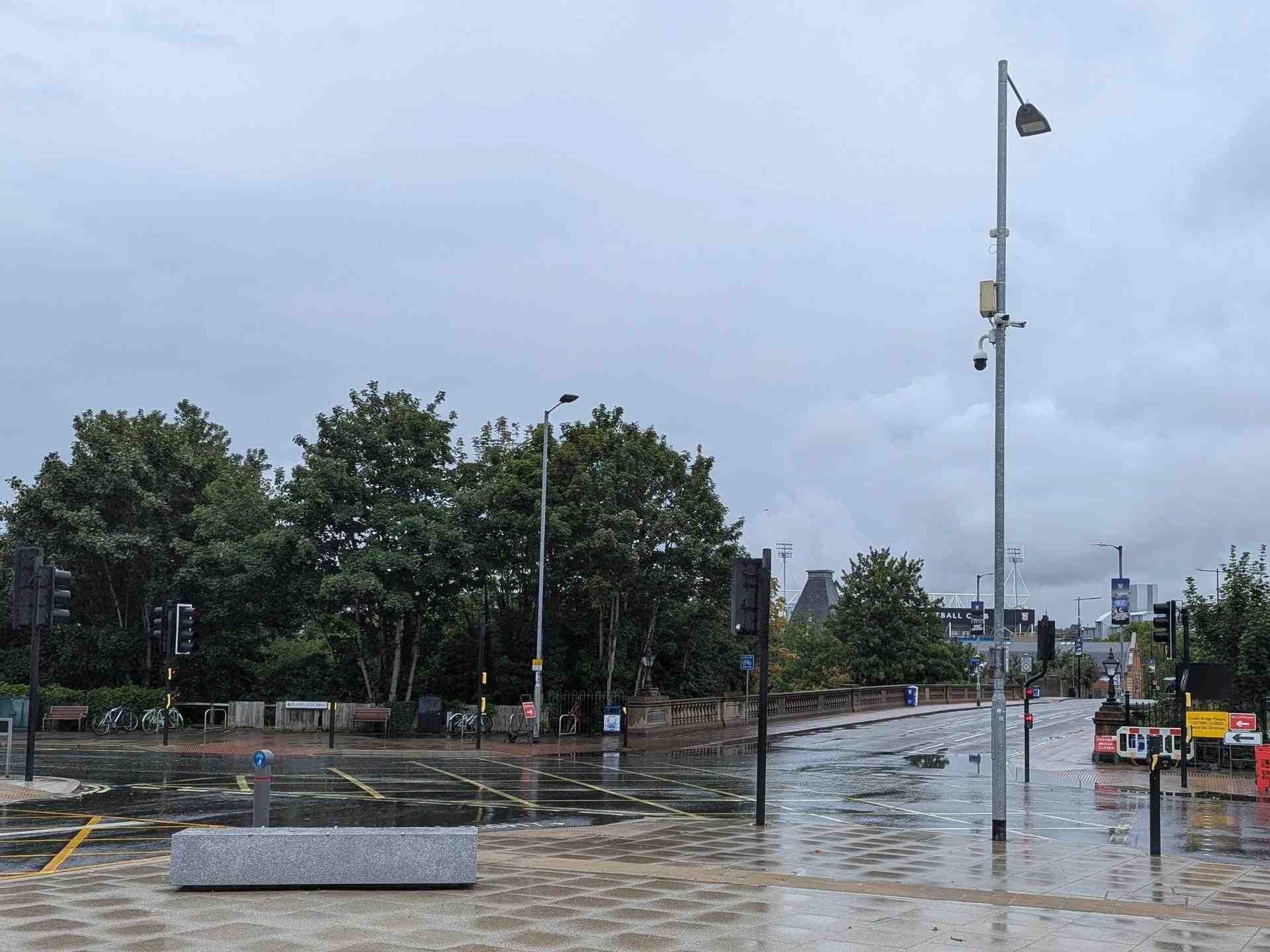 In the foreground, a pedestrian area with a concrete bar to sit on. Behind it, a junction with one one-lane, two three-lane and one four-lane road, controlled by traffic lights. In the distance is the Portman Road football stadium.