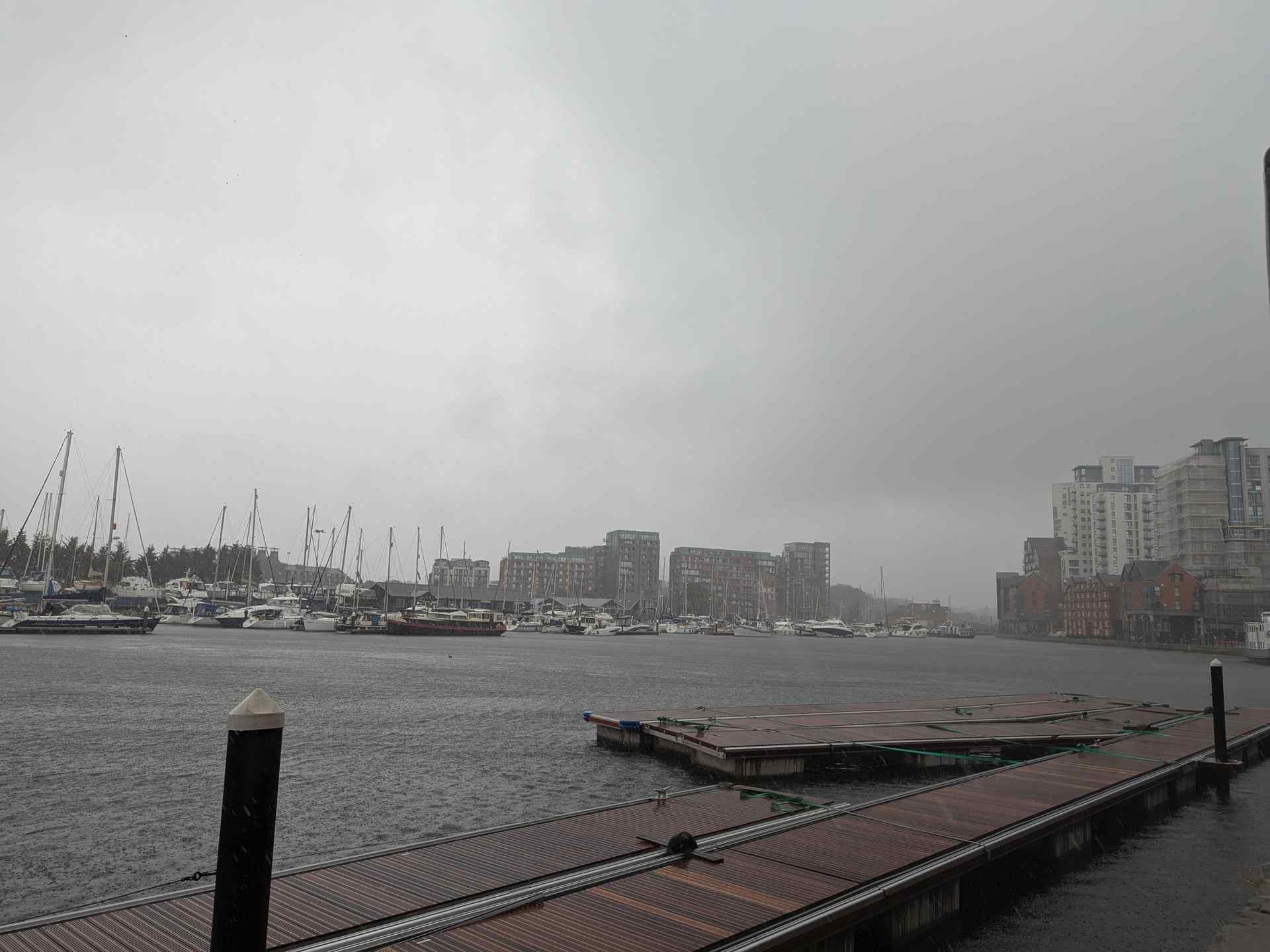 A dull sky atop grey water peppered with raindrops. To the left, there are lots of boats and small yachts moored on the other side of the marina. Centre and right, high-rise residential buildings. In the foreground, disused floating walkways formerly used by the marina.